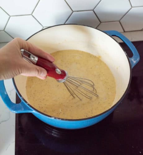 A hand uses a whisk to stir a creamy sauce in a blue pot on a stovetop, with a white hexagon-tiled backsplash visible in the background.