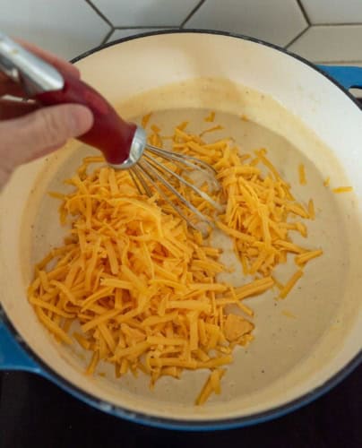 A hand uses a whisk to stir shredded cheddar cheese into a creamy sauce in a blue pot on a stove, with white tile backsplash in the background.