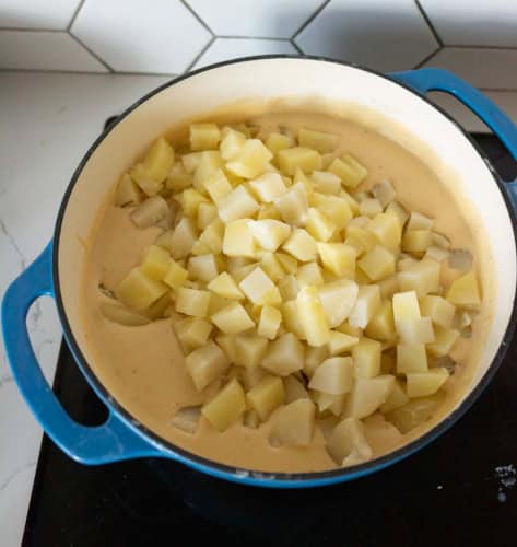 A blue pot filled with creamy sauce and topped with a layer of diced potatoes sits on a black stovetop in a kitchen with white hexagon tile backsplash.