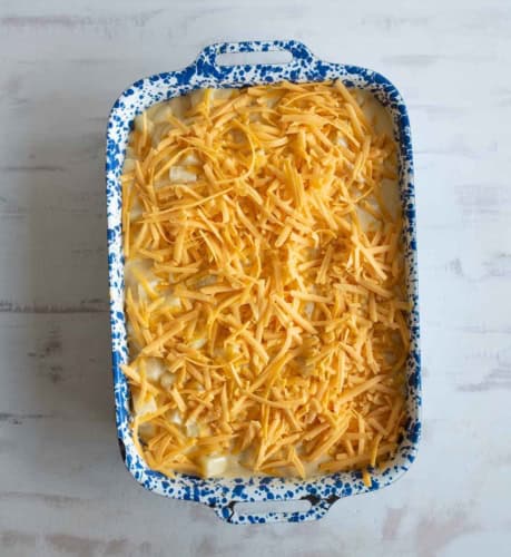 A blue and white speckled baking dish filled with a mixture topped with a generous layer of shredded cheddar cheese, ready to be baked. The dish is on a light-colored surface.