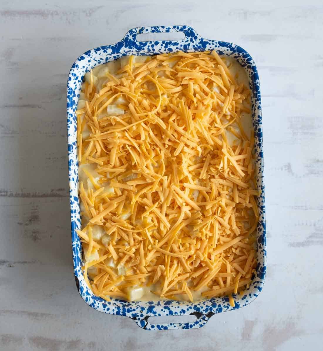 A blue and white speckled baking dish filled with a mixture topped with a generous layer of shredded cheddar cheese, ready to be baked. The dish is on a light-colored surface.