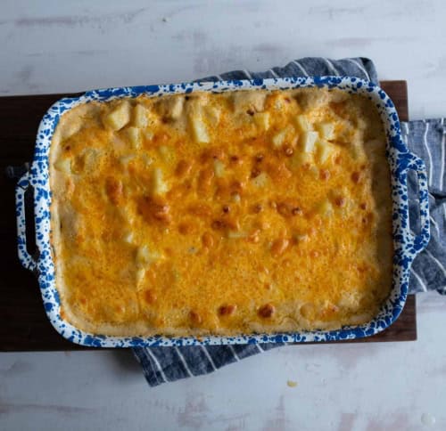 A blue and white speckled baking dish filled with cheesy, golden-brown scalloped potatoes sits on a striped kitchen towel and wooden board.
