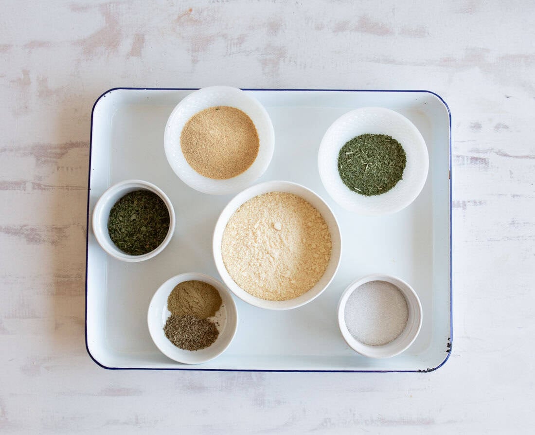 A white tray holds six small white bowls filled with various dried herbs and powders, arranged neatly on a light-colored surface.
