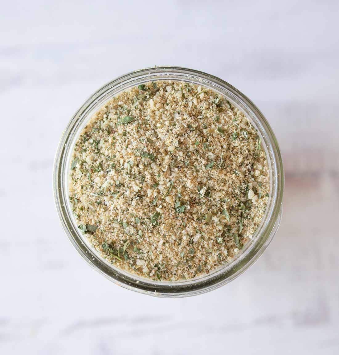 A glass jar filled with a coarse, mixed seasoning blend containing green and beige specks, viewed from above on a light, blurred surface.