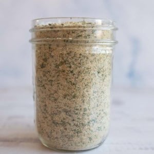 A glass jar filled with garlic parmesan seasoning, showing a blend of fine crumbs and green herbs, placed on a light-colored surface with a soft, blurred background.