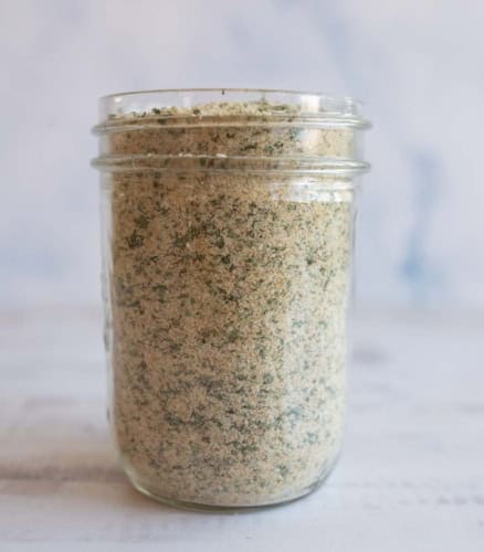 A glass jar filled with garlic parmesan seasoning, showing a blend of fine crumbs and green herbs, placed on a light-colored surface with a soft, blurred background.