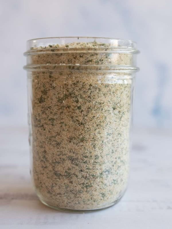 A glass jar filled with garlic parmesan seasoning, showing a blend of fine crumbs and green herbs, placed on a light-colored surface with a soft, blurred background.