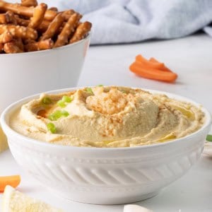 A white bowl of garlic hummus recipe without tahini, garnished with chopped green onions, surrounded by lemon wedges, garlic cloves, carrot sticks, and a bowl of pretzel rods, set on a light countertop with a blue cloth in the background.