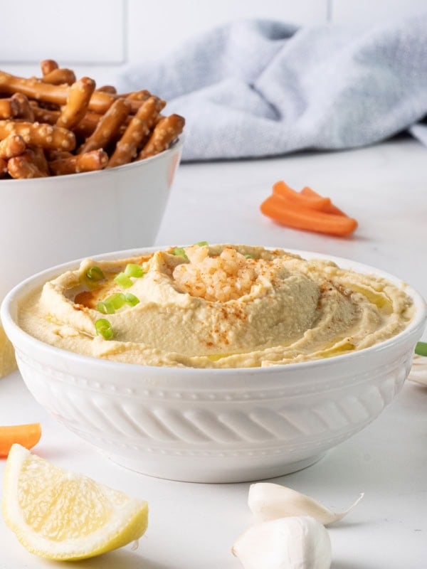 A white bowl of garlic hummus recipe without tahini, garnished with chopped green onions, surrounded by lemon wedges, garlic cloves, carrot sticks, and a bowl of pretzel rods, set on a light countertop with a blue cloth in the background.