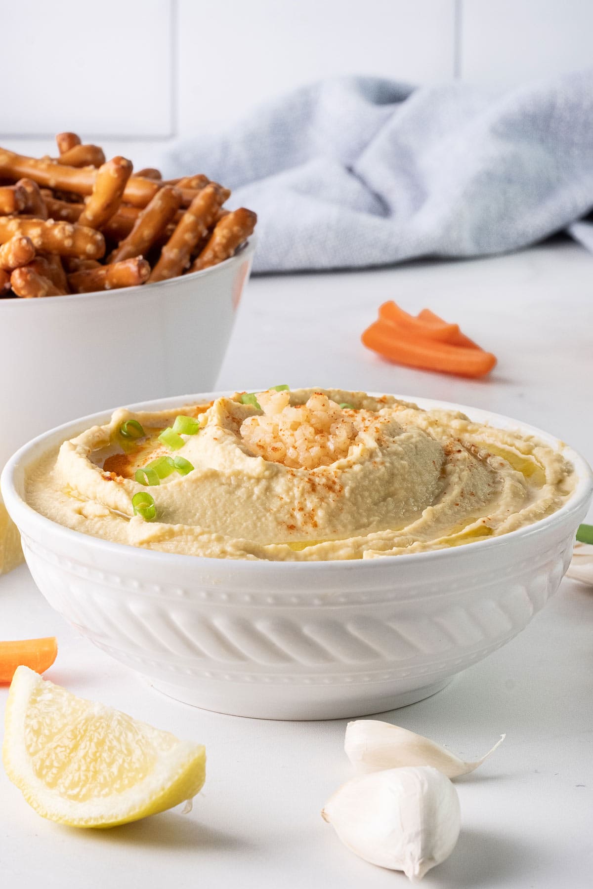 A white bowl of garlic hummus recipe without tahini, garnished with chopped green onions, surrounded by lemon wedges, garlic cloves, carrot sticks, and a bowl of pretzel rods, set on a light countertop with a blue cloth in the background.