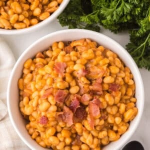 A white bowl filled with homemade baked beans and pieces of bacon, placed on a white surface next to fresh parsley, a white cloth, black spoons, and part of another bowl of beans.
