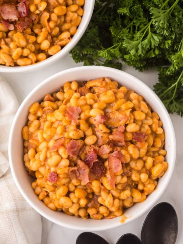 A white bowl filled with homemade baked beans and pieces of bacon, placed on a white surface next to fresh parsley, a white cloth, black spoons, and part of another bowl of beans.