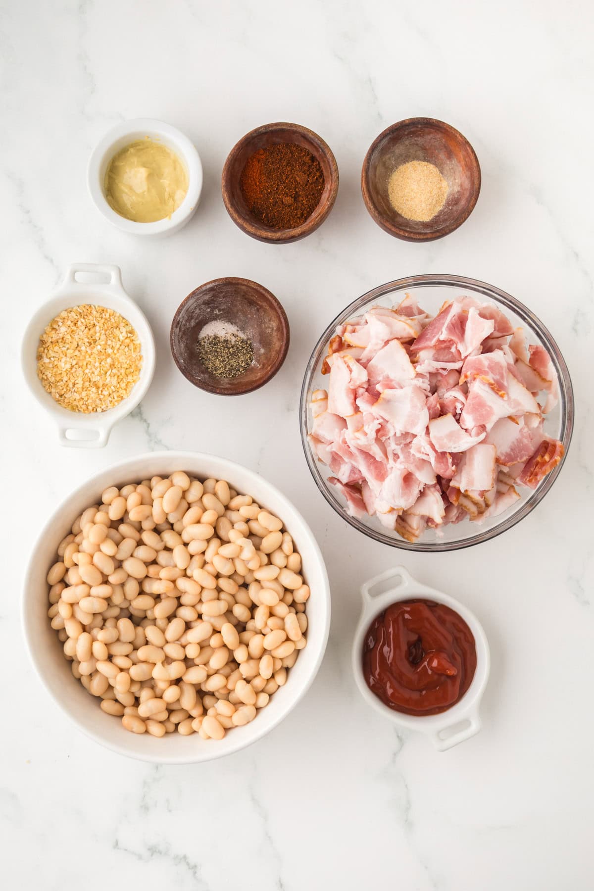 Ingredients for homemade baked beans arranged on a white marble surface, including bowls of white beans, chopped bacon, ketchup, mustard, dried minced onion, black pepper, chili powder, and garlic powder.