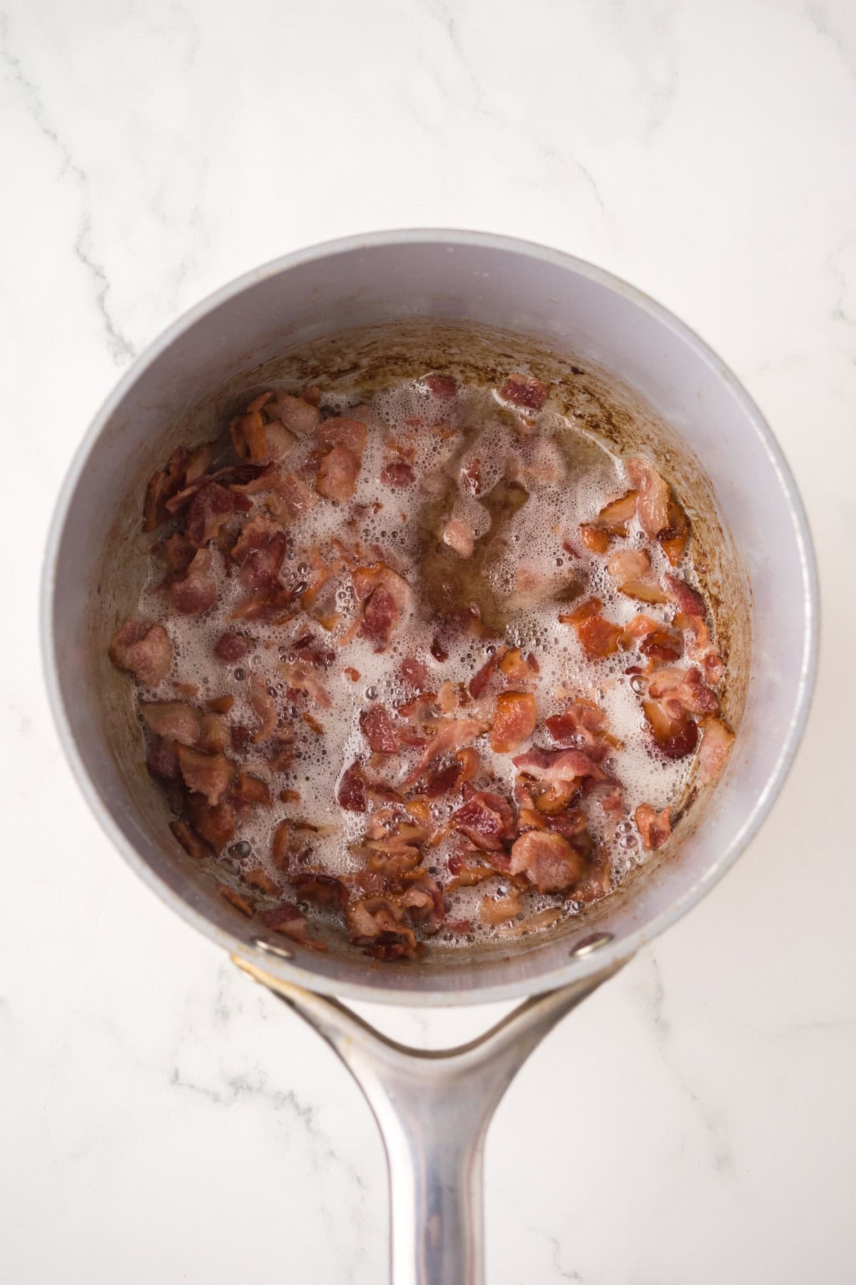 A saucepan containing chopped bacon pieces sizzling and bubbling in their own grease on a white marble surface, the perfect base for homemade baked beans.