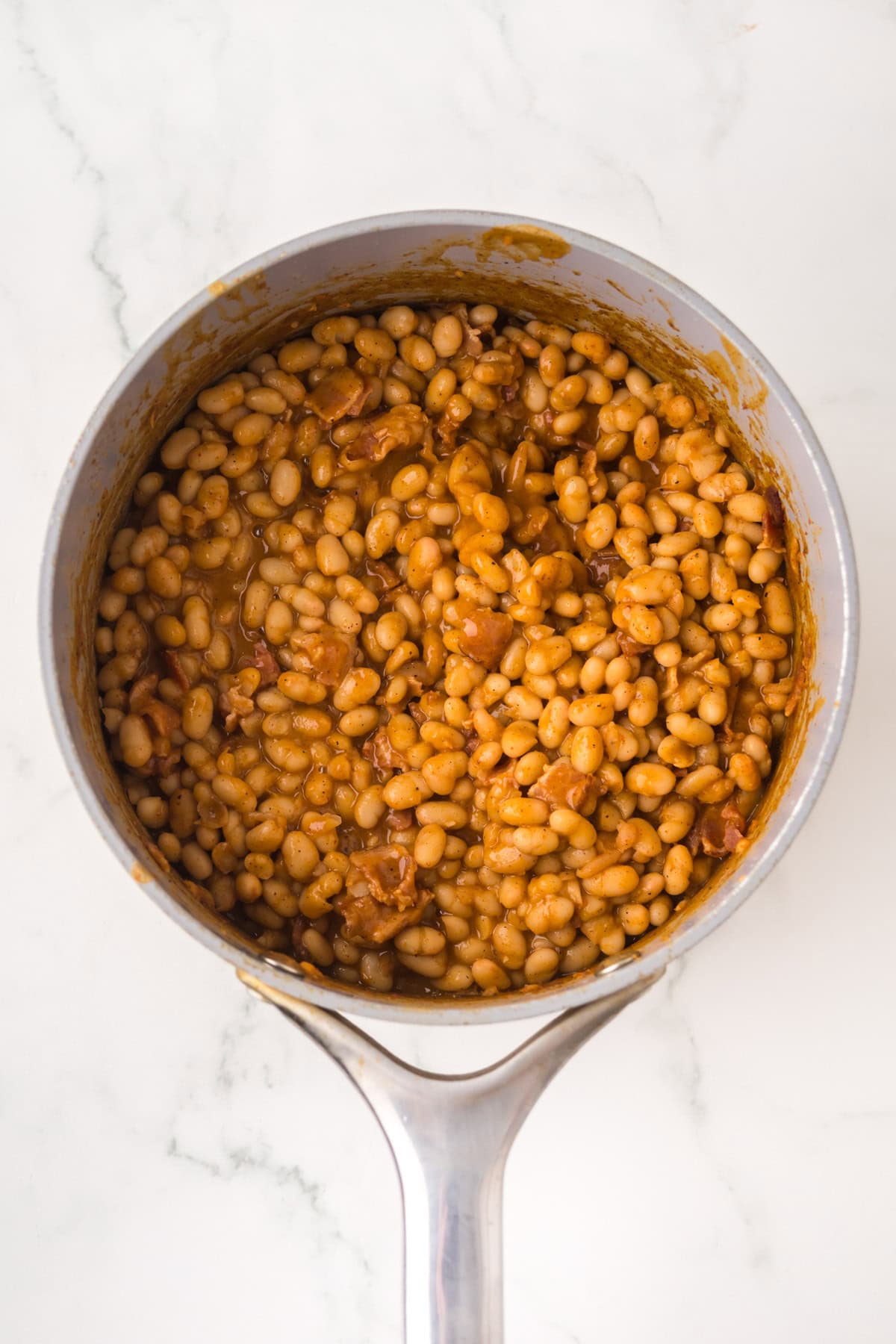 A saucepan filled with homemade baked beans in a thick, reddish-brown sauce, placed on a white marble surface.