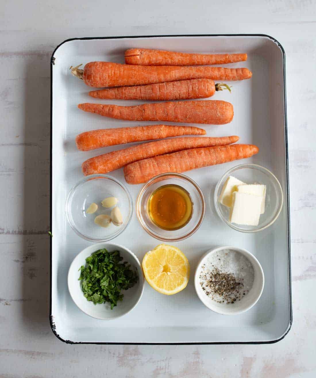 A white tray holding whole carrots, three garlic cloves, a small glass bowl of honey, a pat of butter, a dish of chopped parsley, half a lemon, and a dish of salt and pepper on a light surface.