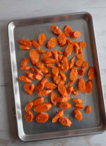 Sliced carrots coated with seasoning are spread out on a metal baking sheet, ready to be roasted. The baking sheet sits on a light-colored surface.