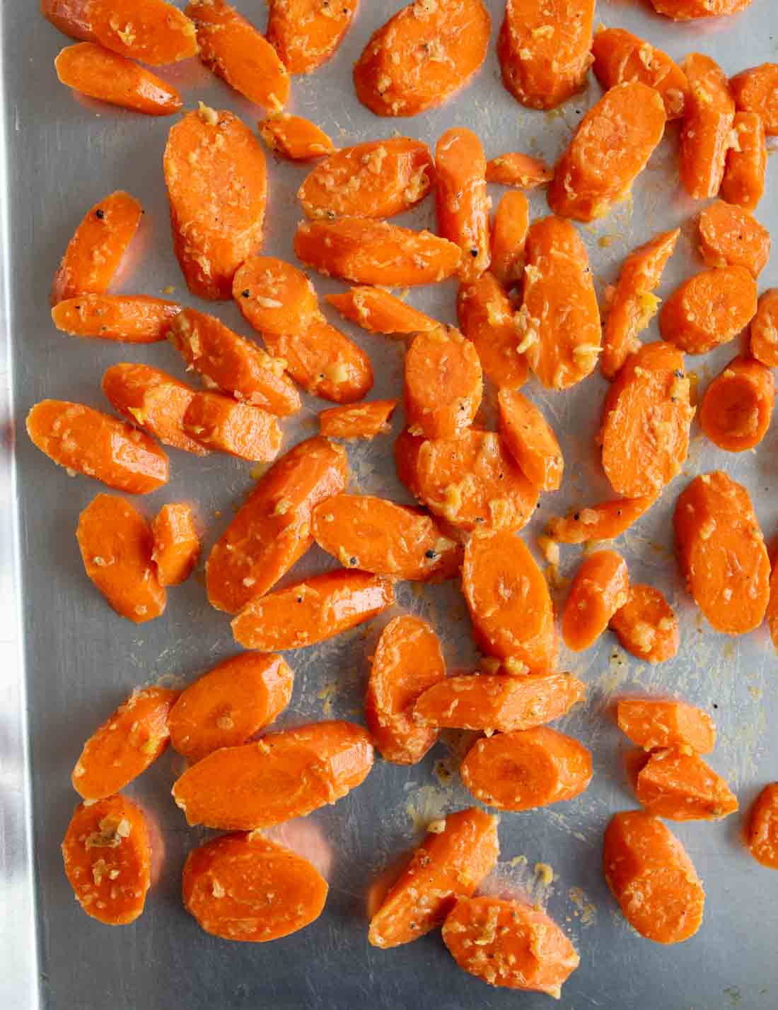 Sliced carrots coated in seasoning are spread out on a metal baking sheet, ready for roasting.