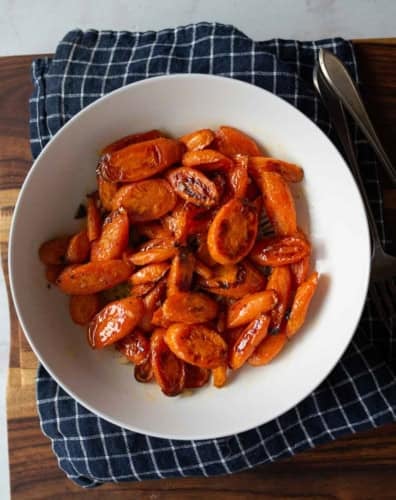 A white bowl filled with roasted, sliced carrots sits on a dark blue checkered cloth, placed on a wooden surface with a fork and knife nearby.