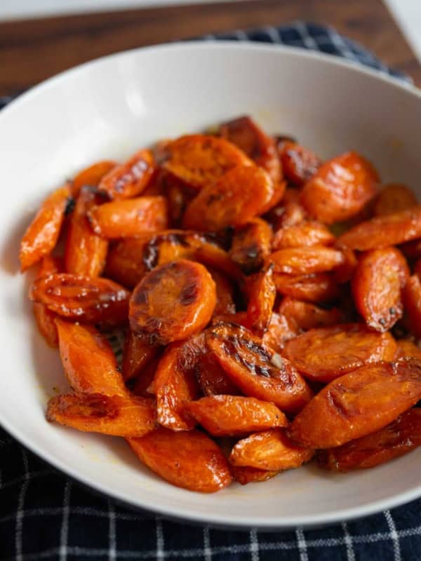 A white bowl filled with honey roasted carrots sits on a dark, checkered cloth. The carrots are glazed and slightly caramelized, giving them a shiny, golden-brown appearance.