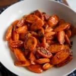 A white bowl filled with roasted, sliced carrots with a caramelized glaze, placed on a dark checkered cloth.