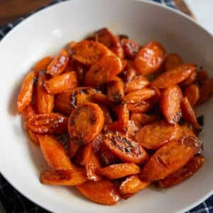 A white bowl filled with roasted, sliced carrots with a caramelized glaze, placed on a dark checkered cloth.