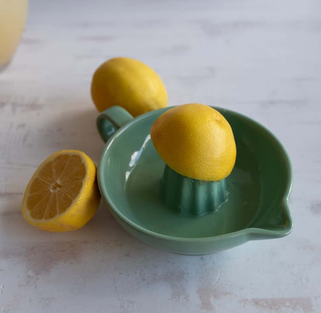 A green ceramic citrus juicer holds half a lemon, with another whole lemon and a lemon half resting on a light-colored surface nearby.
