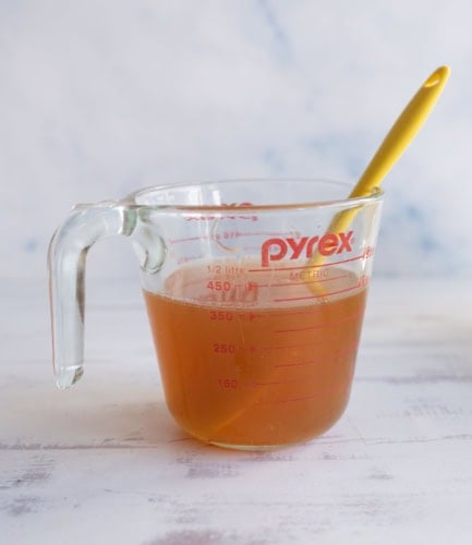 A glass Pyrex measuring cup with brown liquid inside sits on a light surface. A yellow spoon is resting in the cup. The cup has red measurement markings on the side.