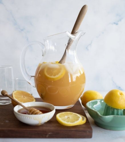 A glass pitcher of iced lemon tea with lemon slices and a wooden spoon sits on a table, surrounded by two empty glasses, fresh lemons, a lemon squeezer, and a bowl of honey with a honey dipper.
