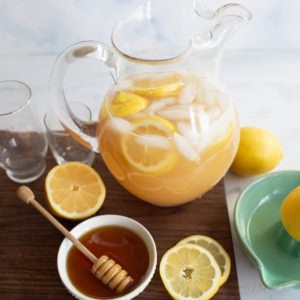 A glass pitcher of iced lemonade with lemon slices sits on a wooden board, next to two empty glasses, a bowl of honey with a dipper, and sliced lemons. A green dish holds a whole lemon nearby.