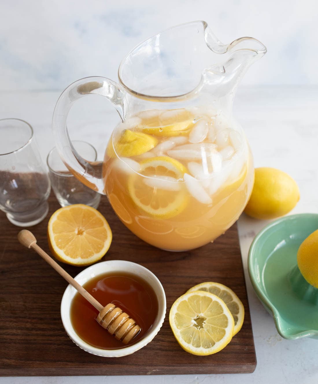 A glass pitcher of iced lemonade with lemon slices sits on a wooden board, next to two empty glasses, a bowl of honey with a dipper, and sliced lemons. A green dish holds a whole lemon nearby.