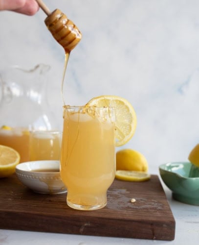 A glass of lemonade garnished with a lemon slice sits on a wooden board. Honey is being drizzled into the glass from a honey dipper, with lemons and a jar of lemonade visible in the background.