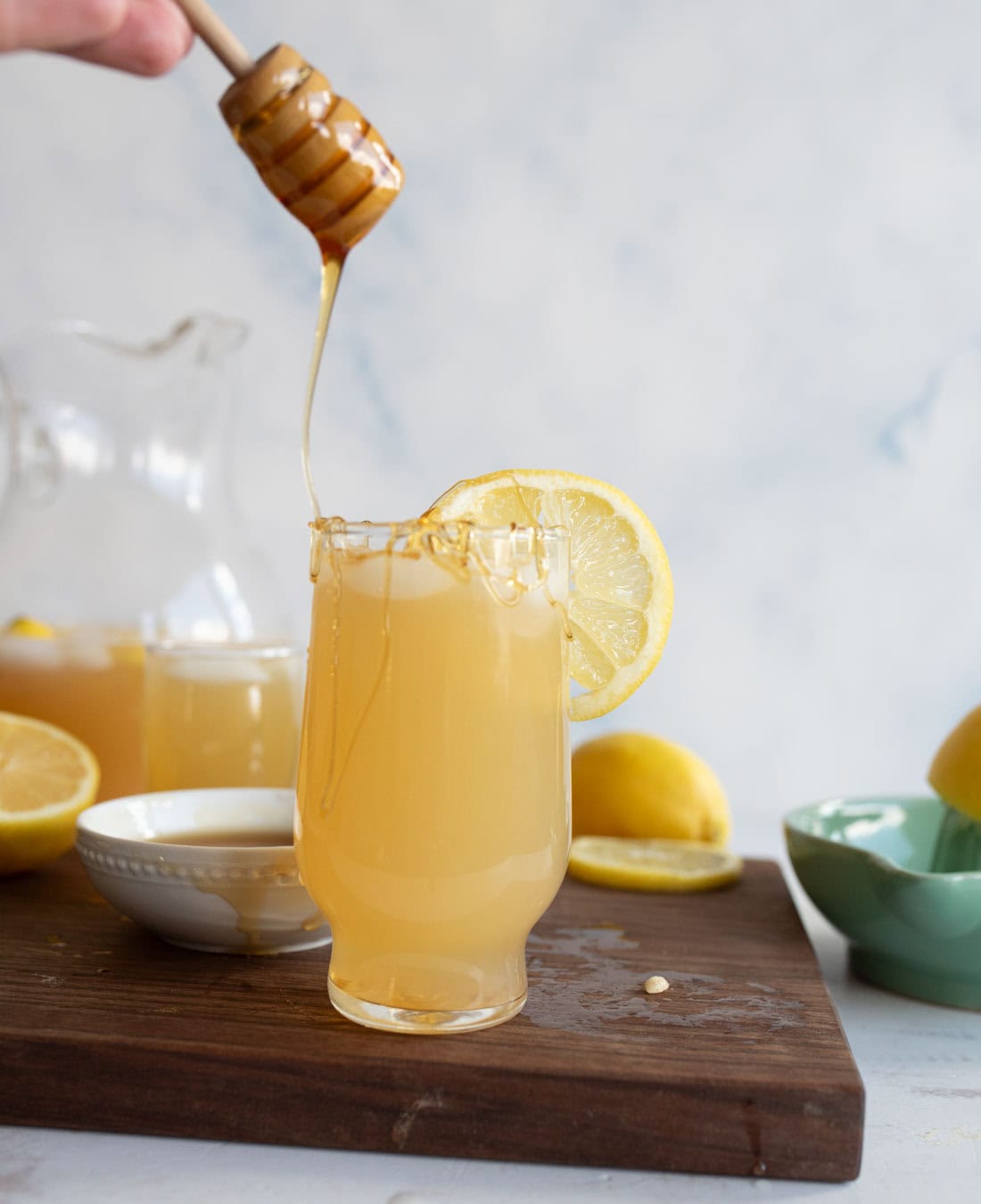 A glass of lemonade garnished with a lemon slice sits on a wooden board. Honey is being drizzled into the glass from a honey dipper, with lemons and a jar of lemonade visible in the background.