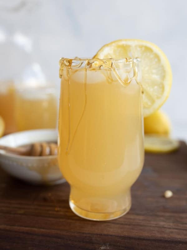 A glass of pale yellow honey lemonade with honey drizzled on the rim and a lemon slice garnish, sits on a wooden surface next to a bowl of honey and more lemon slices in the background.
