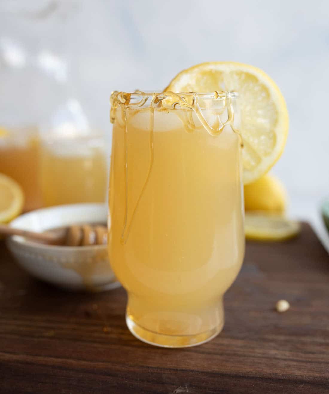A glass of pale yellow honey lemonade with honey drizzled on the rim and a lemon slice garnish, sits on a wooden surface next to a bowl of honey and more lemon slices in the background.