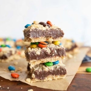 Three dessert bars stacked on top of each other, featuring a crumbly base, a thick chocolate layer, and colorful candy-coated chocolates, with more bars and candies scattered in the background.