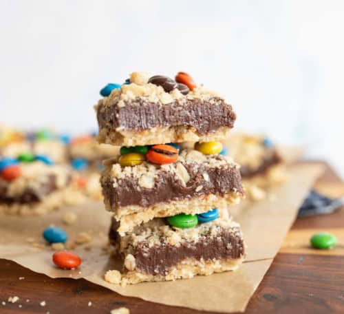 Three dessert bars stacked on top of each other, featuring a crumbly base, a thick chocolate layer, and colorful candy-coated chocolates, with more bars and candies scattered in the background.