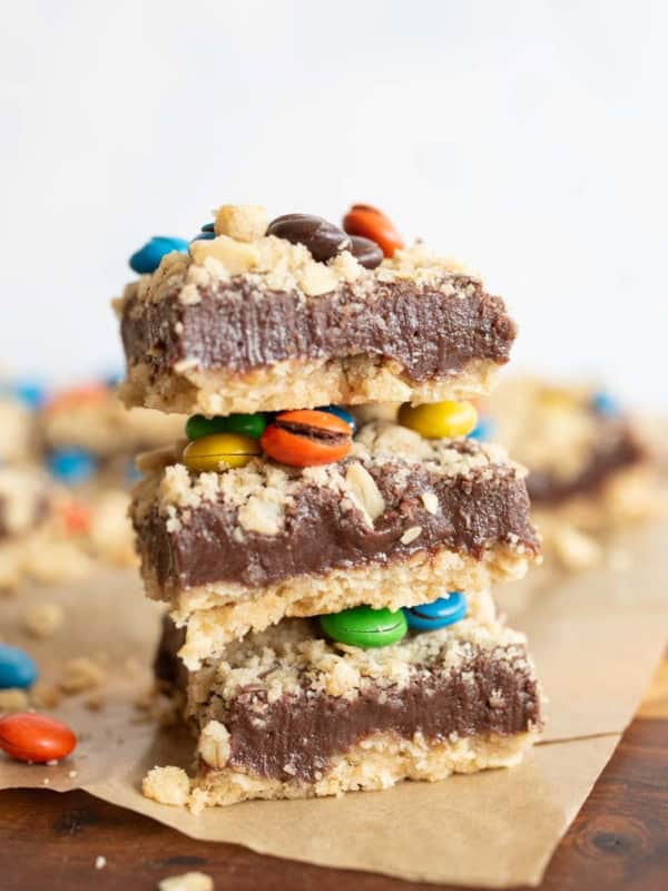 Three dessert bars stacked on top of each other, featuring a crumbly base, a thick chocolate layer, and colorful candy-coated chocolates, with more bars and candies scattered in the background.
