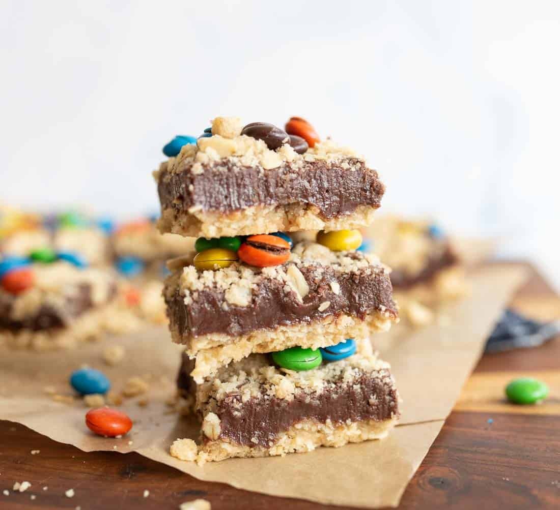Three dessert bars stacked on top of each other, featuring a crumbly base, a thick chocolate layer, and colorful candy-coated chocolates, with more bars and candies scattered in the background.