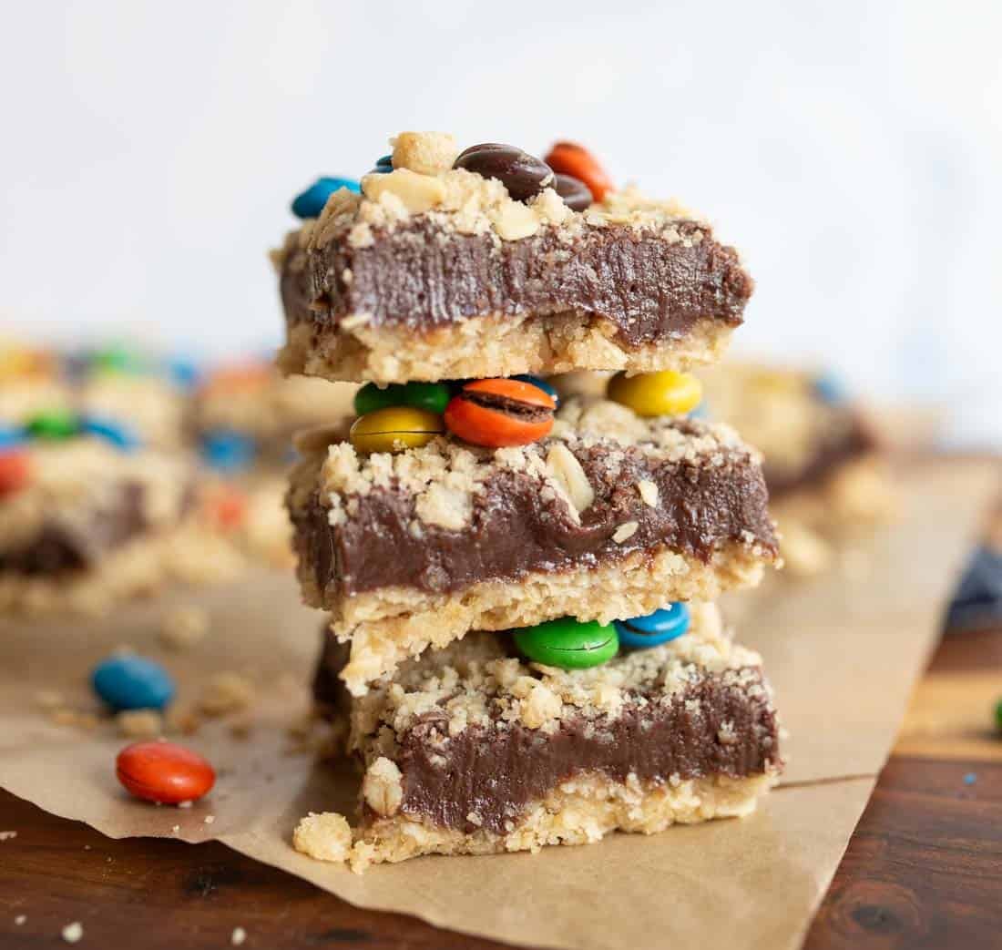 Three chocolate fudge bars with a crumbly topping and colorful candy-coated chocolates are stacked on parchment paper, with more bars and candies blurred in the background.