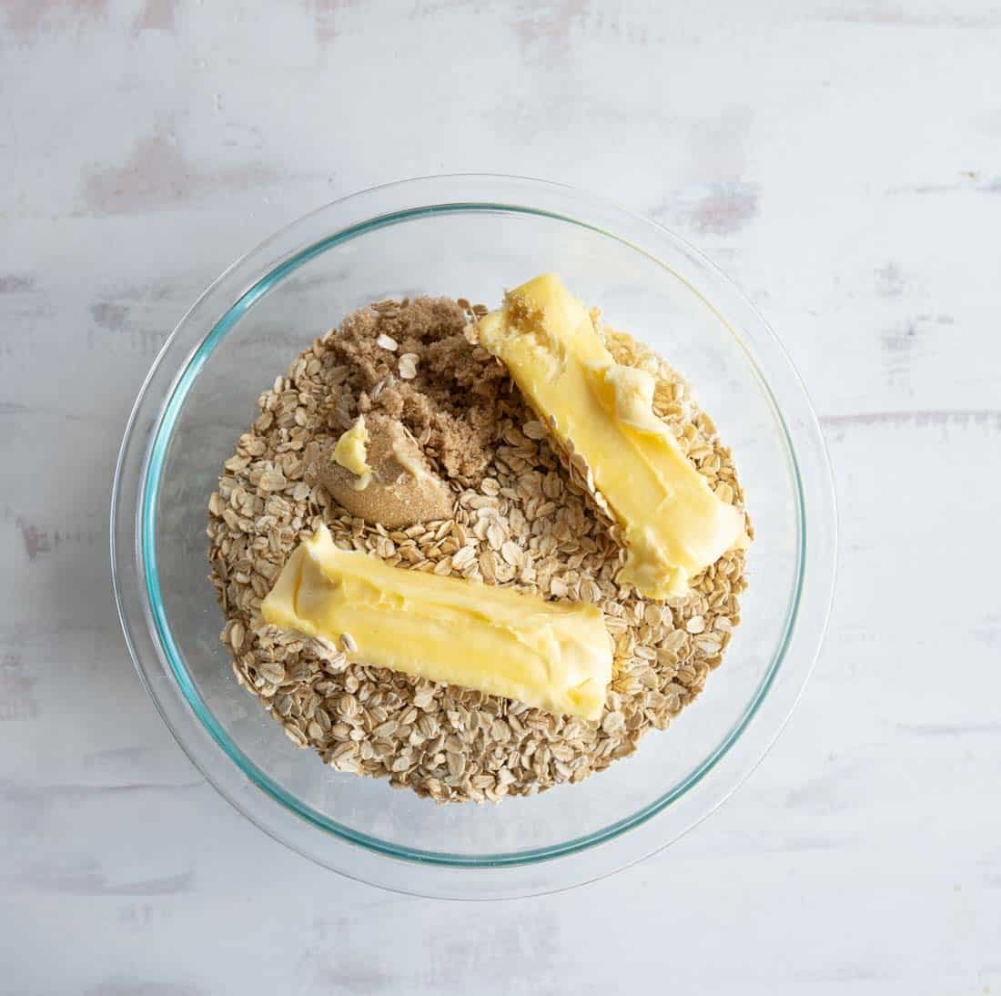 A clear glass bowl containing rolled oats, brown sugar, and two sticks of butter on a white surface.
