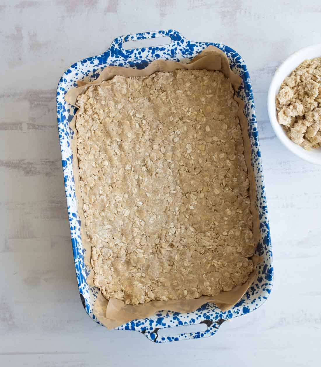 A rectangular oat crumble bar baked in a blue and white speckled baking dish lined with parchment paper, next to a white bowl of oat mixture on a light-colored surface.