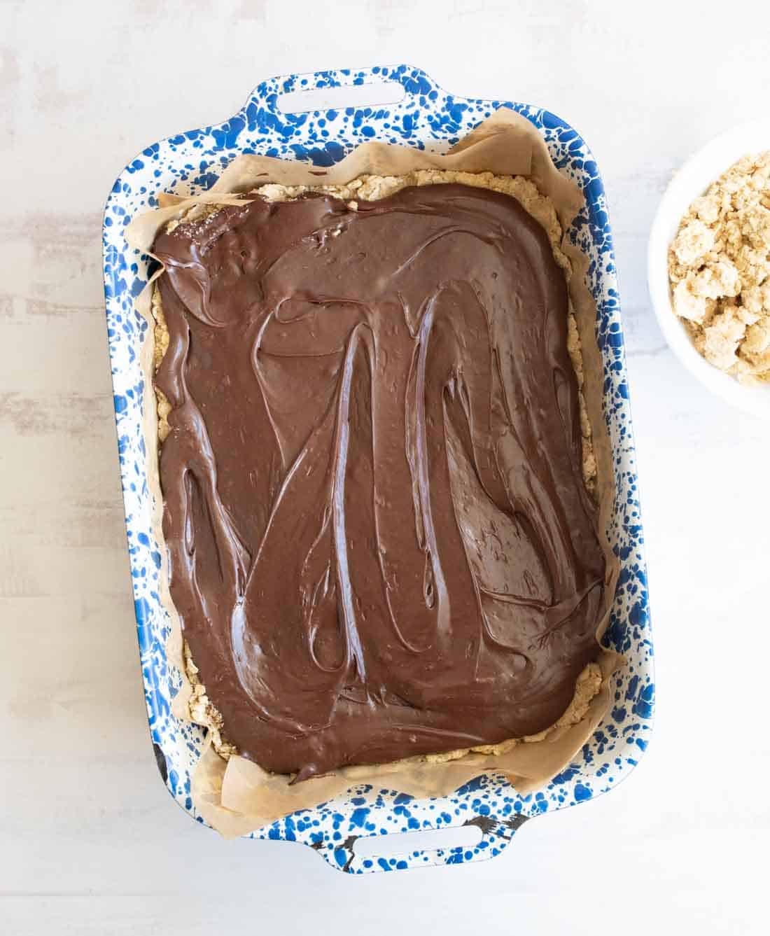 A baking dish lined with parchment paper holds a dessert topped with glossy, swirled chocolate. The dish is blue and white speckled. Next to it is a bowl of crumbled ingredients.