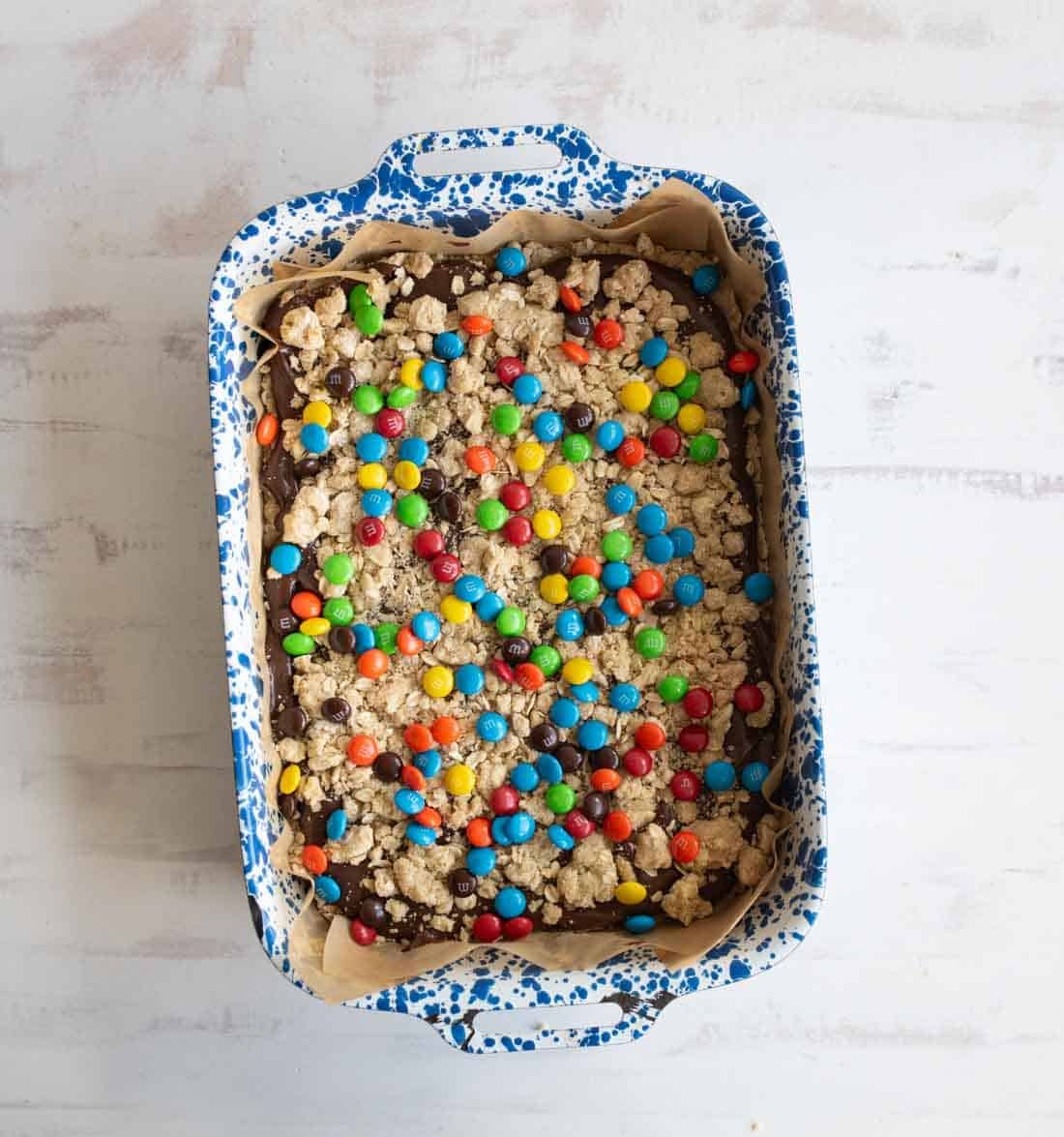 A rectangular blue and white speckled baking dish filled with a crumbly dessert topped with colorful candy-coated chocolates, sitting on a light-colored surface.