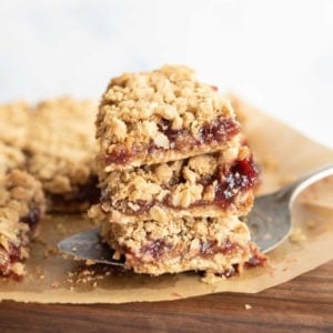 A close-up of three stacked raspberry oatmeal bars on parchment paper, with a metal spatula lifting the stack. The bars have a crumbly oat topping and a layer of red jam in the center.
