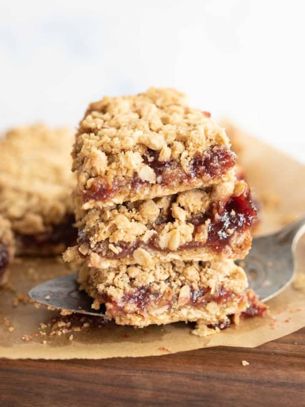 A close-up of three stacked raspberry oatmeal bars on parchment paper, with a metal spatula lifting the stack. The bars have a crumbly oat topping and a layer of red jam in the center.