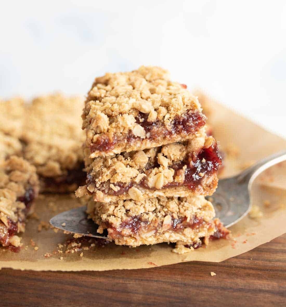 A close-up of three stacked raspberry oatmeal bars on parchment paper, with a metal spatula lifting the stack. The bars have a crumbly oat topping and a layer of red jam in the center.