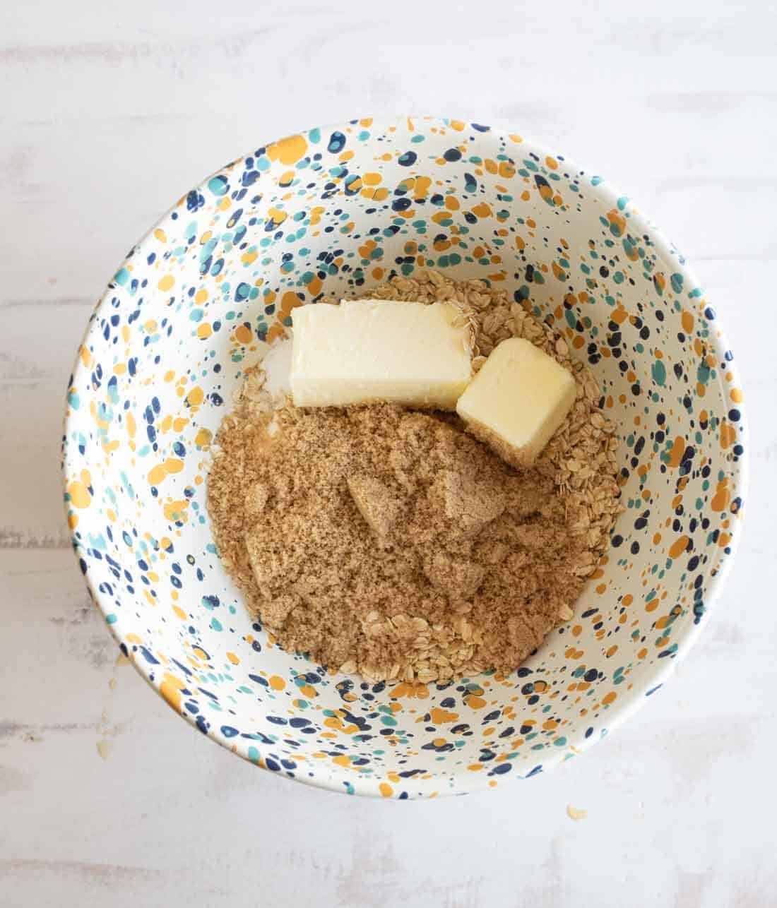 A patterned bowl containing brown sugar, butter, and oats sits on a white surface, ready for mixing.