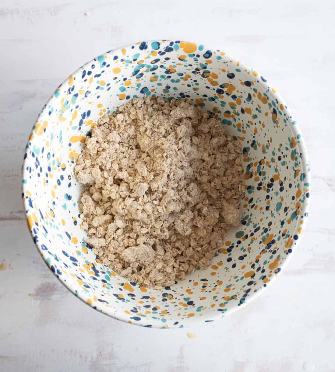 A patterned bowl with blue, yellow, and orange dots holds a mixture of crumbled dry ingredients, possibly oats or streusel, on a light surface.