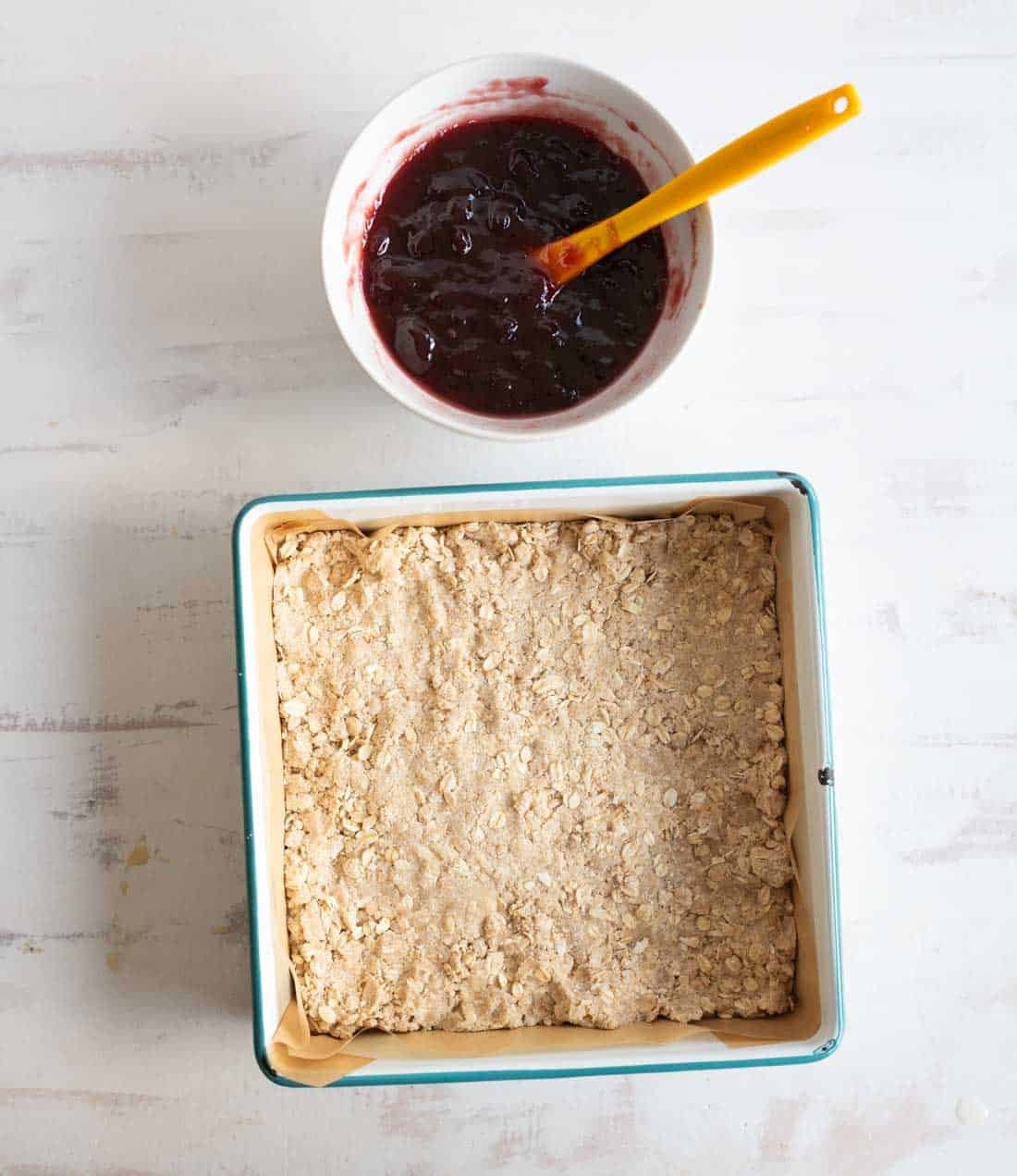 A square baking pan filled with an unbaked oat crust sits on a white surface. Above it is a bowl of dark red fruit jam with a yellow spatula resting inside.